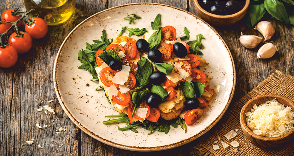 Teller mit Tomaten, schwarzen Oliven, Rucola, Basilikum und Parmesan auf einem rustikalen Holztisch mit Knoblauch, Olivenöl und Parmesan in kleinen Schalen.
